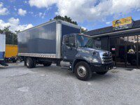 A dark blue box truck is parked outside a building with a sign reading “Box Trucks USA.” The sky is partly cloudy and another yellow truck is partially visible in the background.