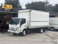 A white Isuzu box truck is parked in front of a building with a sign advertising box truck sales, rentals, and finance. Another white box truck is partially visible on the right. Trees and power lines are in the background.