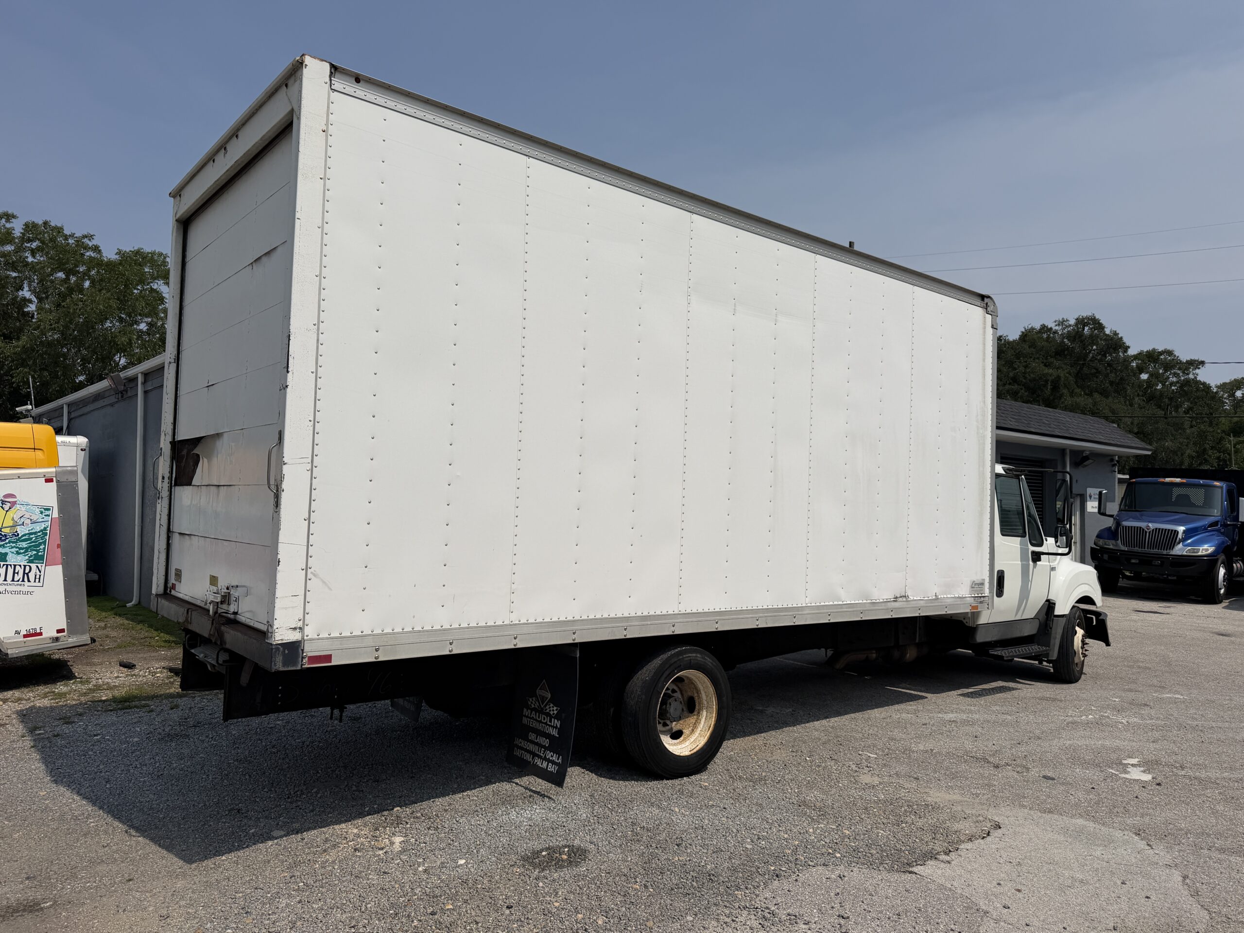 White box truck parked on gravel lot with blue truck in background.